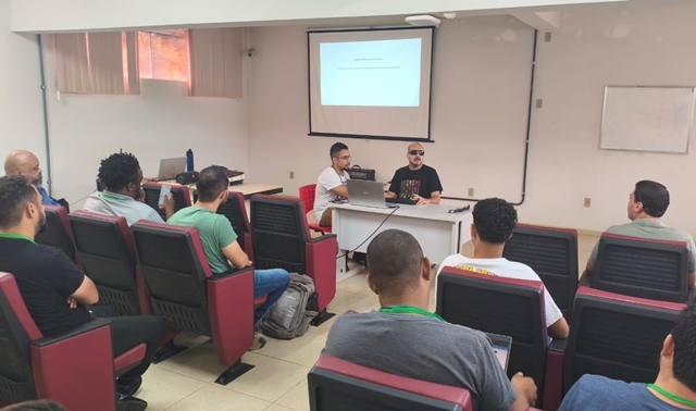 Para cego ver: Sala de aula com cadeiras vermelhas e pretas ocupadas por cerca de dez pessoas. Na frente, duas pessoas estão sentadas atrás de uma mesa branca com notebook. À esquerda, um homem de camiseta clara auxilia na condução da atividade. À direita, o palestrante e ex-aluno Gláucio Brandão, de camiseta preta e óculos escuros, fala ao público. Ao fundo, há uma tela de projeção com slide. Materia_glaucio_2_-_destaque.jpg